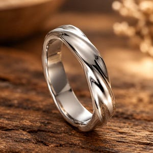 Könnte beinhalten: Ein polierter Ring in Silberoptik mit einem durchgehenden, gedrehten oder gewellten Design entlang des äußeren Bandes. Die Metalloberfläche ist glatt und reflektierend und fängt das Licht ein, um die geschwungenen Kurven hervorzuheben.