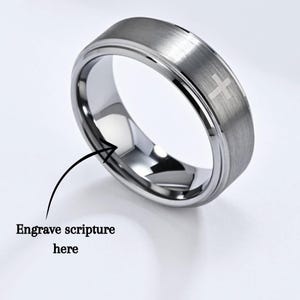 Peut inclure: Une bague en métal argenté avec une finition brossée et un bord biseauté poli. La surface extérieure présente un petit symbole de croix gravé. Une flèche pointe vers l'anneau intérieur lisse avec le texte Engrave scripture here en dessous.