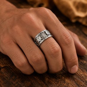 May include: A wide silver-toned metal ring featuring a repeating gear or cogwheel pattern engraved around the entire band. The surface has a brushed finish, and the mechanical design elements are evenly spaced, creating a textured appearance.