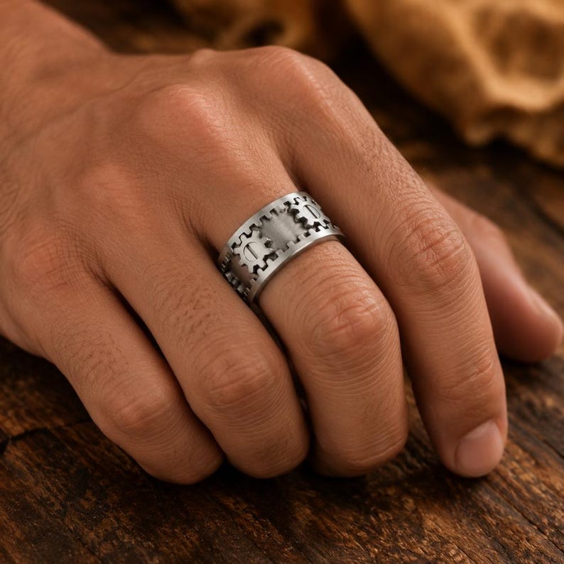 May include: A wide silver-toned metal ring featuring a repeating gear or cogwheel pattern engraved around the entire band. The surface has a brushed finish, and the mechanical design elements are evenly spaced, creating a textured appearance.