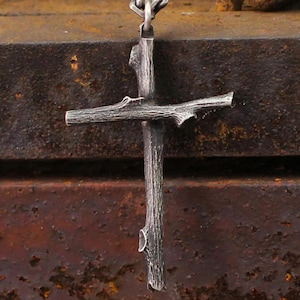 May include: A silver-colored cross pendant with a rustic, twig-like design. The cross is suspended from a chain and features a textured surface, with details mimicking the natural form of branches. The pendant is set against a dark, textured background.