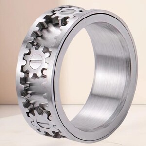 May include: A silver-toned metal ring featuring a wide band with a mechanical design. The outer surface displays a series of raised gear-shaped elements arranged in a row, each with a central horizontal line resembling a screw head detail.