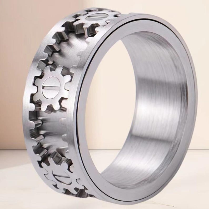 May include: A silver-toned metal ring featuring a wide band with a mechanical design. The outer surface displays a series of raised gear-shaped elements arranged in a row, each with a central horizontal line resembling a screw head detail.