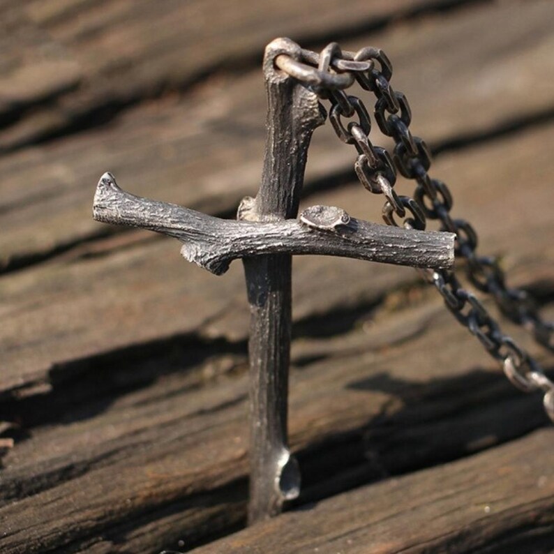 May include: A silver-colored pendant necklace featuring a cross design crafted to resemble a small tree branch. The cross is attached to a chain. The necklace is displayed on a wooden surface.