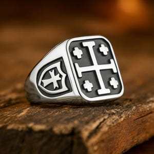 以下が含まれることがあります： 黒い十字のデザインが施された正方形の顔を持つシルバーカラーのリング。リングの側面には盾のエンブレムが付いています。リングは金属製で、木製の表面に表示されています。