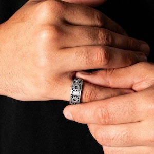May include: A silver-toned metal ring featuring a continuous band design of interlocking gear motifs. The ring has a matte, textured finish with dark recessed areas between the gear teeth, creating a mechanical appearance on the finger.