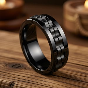 May include: A black ring featuring a central band with a textured, repeating link-style pattern. The ring has a smooth, polished finish on the outer edges and a dark, matte-like appearance on the central decorative section, creating a contrast.