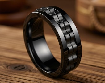 May include: A black ring featuring a central band with a textured, repeating link-style pattern. The ring has a smooth, polished finish on the outer edges and a dark, matte-like appearance on the central decorative section, creating a contrast.