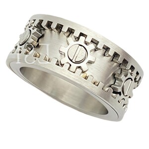 May include: A silver-toned metal ring featuring a brushed finish and a band decorated with raised gear-shaped motifs. The outer edge of the ring has a serrated, cog-like texture, and the surface is accented with small, circular gear elements.