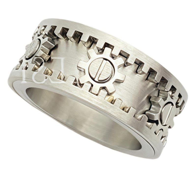 May include: A silver-toned metal ring featuring a brushed finish and a band decorated with raised gear-shaped motifs. The outer edge of the ring has a serrated, cog-like texture, and the surface is accented with small, circular gear elements.