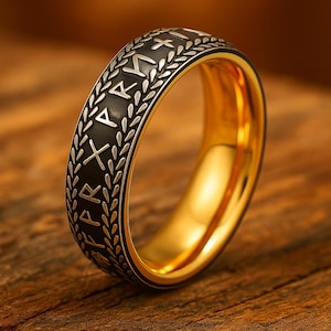 May include: A gold-colored ring with a black band featuring silver-colored runes and a laurel wreath design. The ring has a polished, reflective surface and is set against a wooden background. The ring is a piece of jewelry.