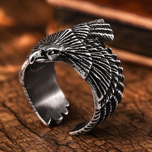 May include: A silver-toned metal ring featuring a detailed eagle design with spread wings that wrap around the band. The surface has a dark, oxidized finish in the recessed areas to highlight the textured feather patterns and the eagle's head.
