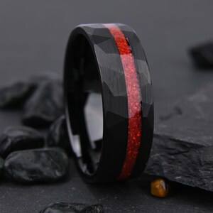 Puede incluir: Un anillo negro con una textura martillada y una incrustación roja vibrante. El anillo tiene un acabado negro brillante y la incrustación roja recorre el centro. El anillo se muestra sobre una superficie oscura con piedras negras.