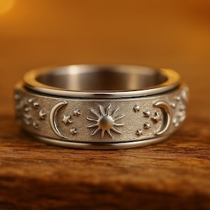 May include: Silver-toned ring featuring a celestial design. The band is adorned with embossed suns, crescent moons, and stars. The ring has a spinning outer band and is set against a warm, wooden background.