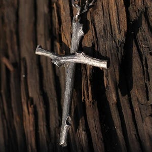 May include: A silver-colored cross pendant with a rustic, twig-like design. The cross is suspended from a chain and features a textured surface, giving it a natural, handcrafted appearance. The pendant is displayed against a dark wood background.