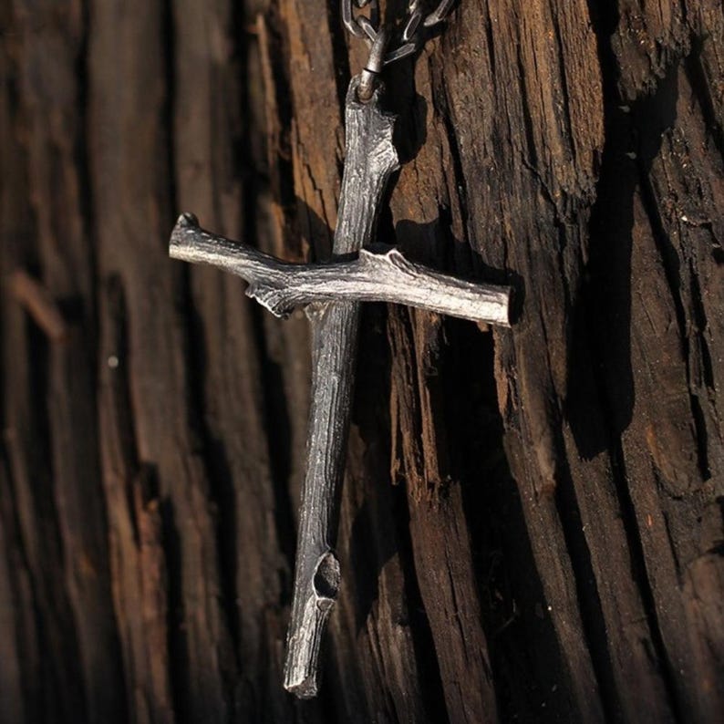 May include: A silver-colored cross pendant with a rustic, twig-like design. The cross is suspended from a chain and features a textured surface, giving it a natural, handcrafted appearance. The pendant is displayed against a dark wood background.