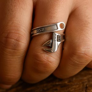 Könnte beinhalten: Ein silberfarbener Ringschlüssel, der sich um einen Finger wickelt. Der Ring hat ein detailliertes Design, mit Zahlen und Markierungen auf dem Metall. Der Ring ist ein einzigartiges Schmuckstück.