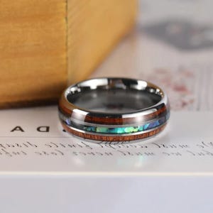 May include: A polished metal ring featuring a central inlay of dark brown wood and a thin, iridescent blue and green crushed stone band running through the middle of the wood, creating a layered, multi-textured appearance on the outer surface.
