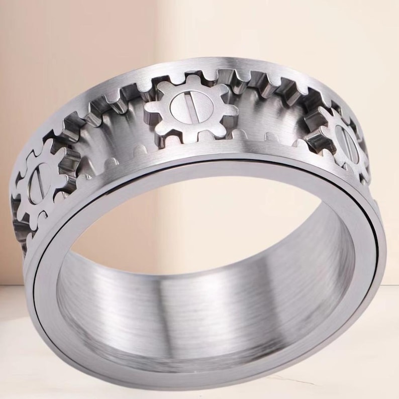 May include: A silver-toned metal ring featuring a rotating outer band design. The surface of the ring is decorated with a series of raised gear-shaped elements, each containing a central horizontal slot resembling a screw head, set against a brushed metal finish.