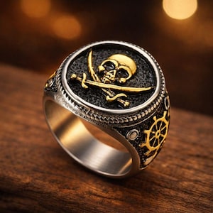 May include: A silver-toned ring with a gold skull and crossed swords design. The ring features a black background with a gold border and a gold ship's wheel on the side. The ring is a bold statement piece.