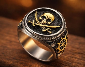 May include: A silver-toned ring featuring a circular face with a gold-colored skull and crossed swords emblem set against a textured black background. The sides of the band are decorated with a gold-colored ship steering wheel motif.