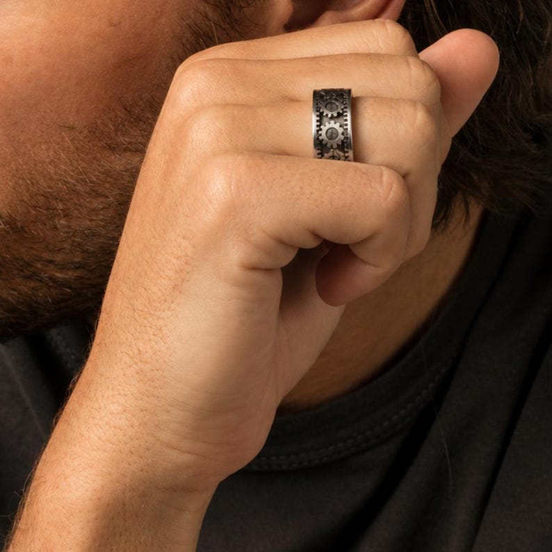 May include: A wide silver-toned metal band ring featuring a repeating mechanical gear pattern around the entire circumference. The ring has a dark, oxidized finish within the recessed areas to highlight the intricate details of the gear teeth.