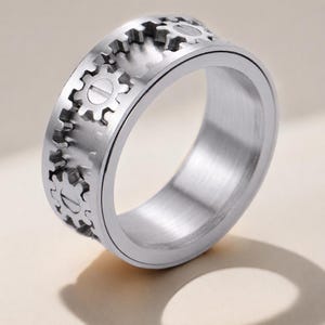 May include: A silver-toned metal ring featuring a band with a series of raised gear-shaped motifs around the exterior. The surface of the ring has a brushed metallic finish, and each gear element includes a central horizontal line detail.