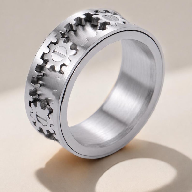 May include: A silver-toned metal ring featuring a band with a series of raised gear-shaped motifs around the exterior. The surface of the ring has a brushed metallic finish, and each gear element includes a central horizontal line detail.