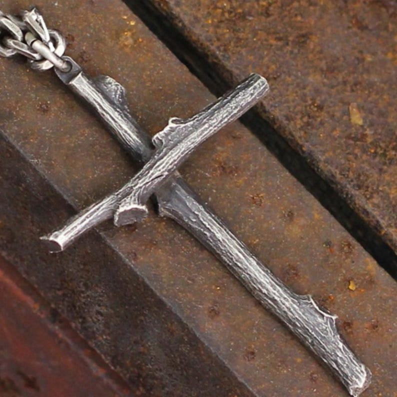 May include: A silver-colored pendant necklace featuring a cross design made to resemble intertwined branches. The pendant is attached to a silver chain. The necklace has a rustic, natural aesthetic.