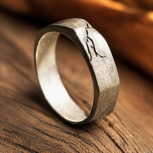 May include: A silver-toned ring with a textured, matte finish and a faceted, angular band design. The surface of the ring features a thin, dark, etched line pattern that resembles a branching or cracked detail on the outer edge of the metal band.