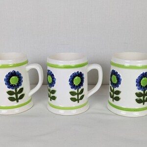 Puede incluir: Tres tazas de cer&aacute;mica blanca con borde y asa verdes, cada una decorada con un dise&ntilde;o floral azul y verde. Las tazas tienen forma cil&iacute;ndrica y un asa robusta.