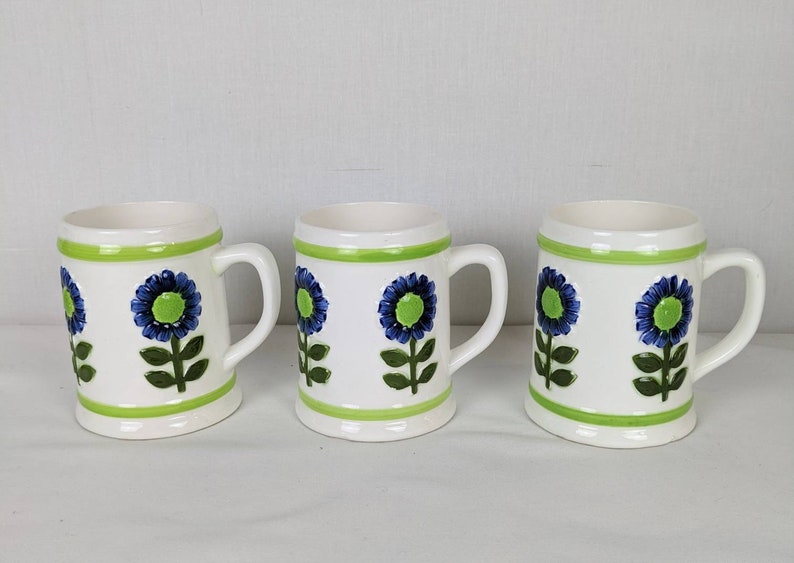 Puede incluir: Tres tazas de cer&aacute;mica blanca con borde y asa verdes, cada una decorada con un dise&ntilde;o floral azul y verde. Las tazas tienen forma cil&iacute;ndrica y un asa robusta.