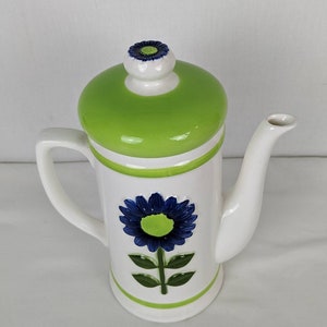 Puede incluir: Tetera de cer&aacute;mica blanca con tapa verde y dise&ntilde;o de flor azul. La tetera tiene asa y boquilla. La flor est&aacute; en el centro de la tetera. La tapa tiene una peque&ntilde;a flor encima.