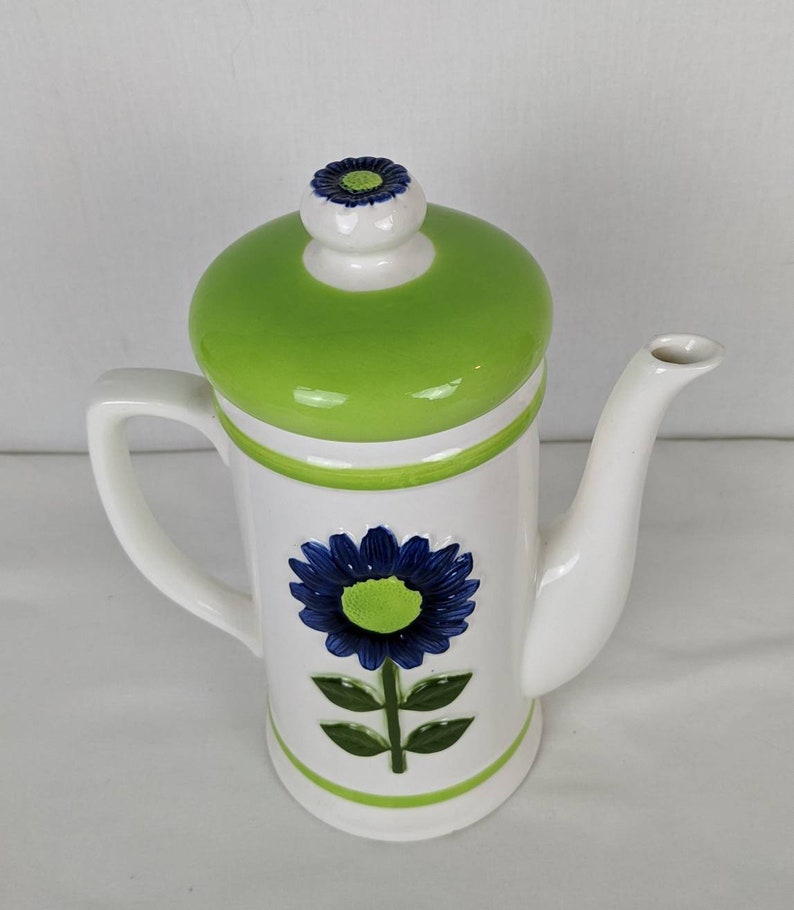 Puede incluir: Tetera de cer&aacute;mica blanca con tapa verde y dise&ntilde;o de flor azul. La tetera tiene asa y boquilla. La flor est&aacute; en el centro de la tetera. La tapa tiene una peque&ntilde;a flor encima.