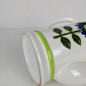 Puede incluir: Jarra de cer&aacute;mica blanca con asa, decorada con una franja verde y motivos florales azules. La jarra tiene un acabado brillante y un dise&ntilde;o cl&aacute;sico, ideal para servir bebidas o como pieza decorativa.