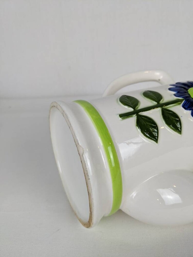 Puede incluir: Jarra de cer&aacute;mica blanca con asa, decorada con una franja verde y motivos florales azules. La jarra tiene un acabado brillante y un dise&ntilde;o cl&aacute;sico, ideal para servir bebidas o como pieza decorativa.