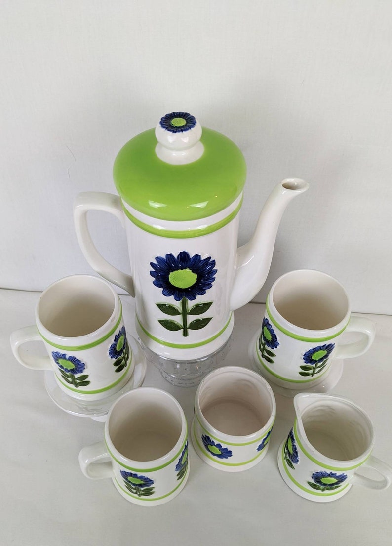 Puede incluir: Juego de caf&eacute; de cer&aacute;mica vintage en blanco y verde, decorado con motivos florales azules. El juego incluye una cafetera, seis tazas y una jarrita para la crema. La cafetera tiene una tapa verde y un dise&ntilde;o floral azul.
