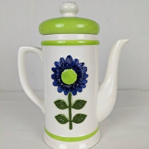 Puede incluir: Cafetera de cer&aacute;mica blanca con un dise&ntilde;o floral azul y verde. La cafetera tiene una tapa y detalles verdes, un asa curva y un pico. El dise&ntilde;o floral incluye una flor azul con centro verde y hojas verdes.