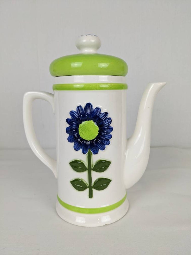 Puede incluir: Cafetera de cer&aacute;mica blanca con un dise&ntilde;o floral azul y verde. La cafetera tiene una tapa y detalles verdes, un asa curva y un pico. El dise&ntilde;o floral incluye una flor azul con centro verde y hojas verdes.