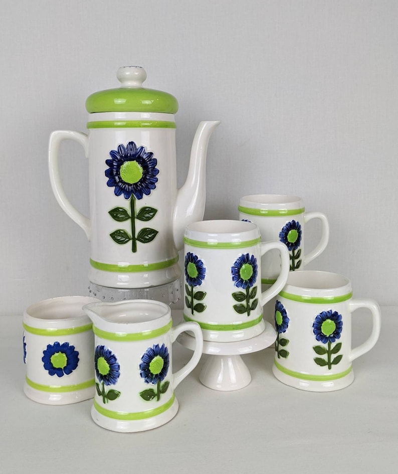 Puede incluir: Un juego de caf&eacute; de cer&aacute;mica blanca con un dise&ntilde;o floral verde y azul. El juego incluye una cafetera, una jarra de crema, un azucarero y cuatro tazas. La cafetera tiene una tapa verde y una gran flor azul.