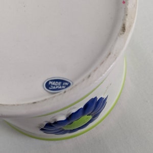 Puede incluir: Contenedor de cer&aacute;mica blanca con un dise&ntilde;o floral azul y verde. El contenedor tiene un borde verde y una flor azul con un centro verde. Es visible una pegatina azul y blanca que dice "MADE IN JAPAN".