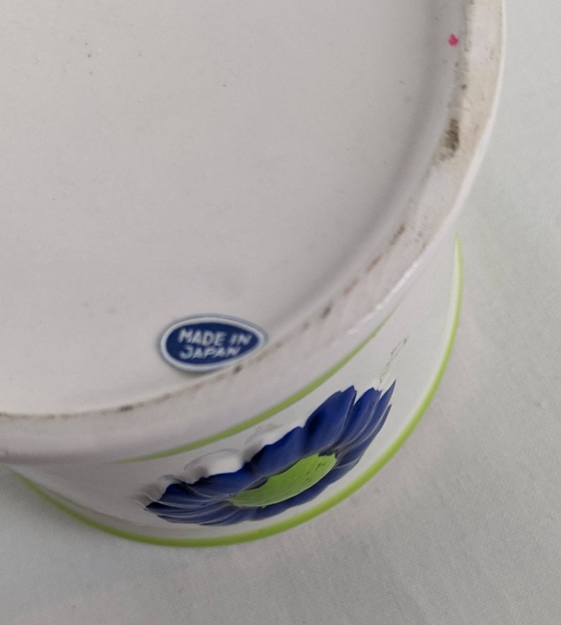 Puede incluir: Contenedor de cer&aacute;mica blanca con un dise&ntilde;o floral azul y verde. El contenedor tiene un borde verde y una flor azul con un centro verde. Es visible una pegatina azul y blanca que dice "MADE IN JAPAN".