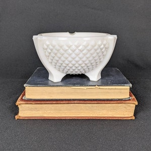 Könnte beinhalten: Weißer Milchglas-Aschenbecher mit Diamantenmuster. Der Aschenbecher steht auf drei Büchern.
