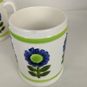 Puede incluir: Taza de cer&aacute;mica blanca con flor azul y detalles verdes. La taza presenta un dise&ntilde;o floral en relieve con una flor azul y centro verde, hojas verdes y bandas verdes en la parte superior e inferior.