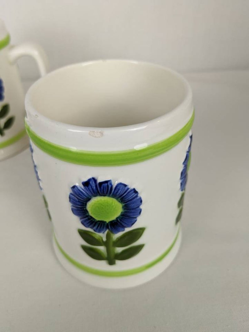 Puede incluir: Taza de cer&aacute;mica blanca con flor azul y detalles verdes. La taza presenta un dise&ntilde;o floral en relieve con una flor azul y centro verde, hojas verdes y bandas verdes en la parte superior e inferior.