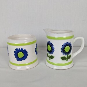 Puede incluir: Un juego de jarra de crema y azucarero de cer&aacute;mica blanca. El juego presenta un dise&ntilde;o floral con detalles en azul y verde. La jarra de crema tiene asa y boquilla. El azucarero es cil&iacute;ndrico. Ambas piezas tienen bandas verdes.