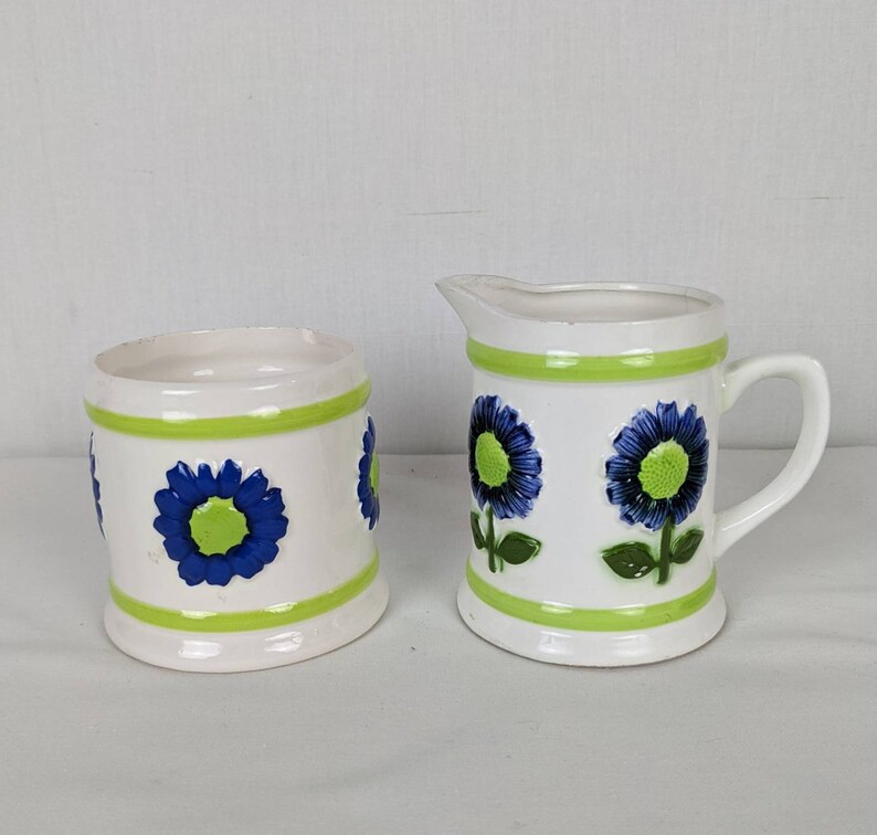 Puede incluir: Un juego de jarra de crema y azucarero de cer&aacute;mica blanca. El juego presenta un dise&ntilde;o floral con detalles en azul y verde. La jarra de crema tiene asa y boquilla. El azucarero es cil&iacute;ndrico. Ambas piezas tienen bandas verdes.