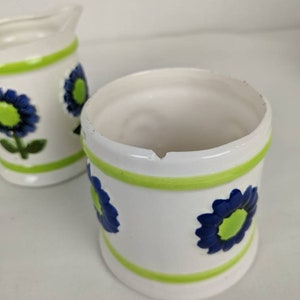 Puede incluir: Un juego de jarra de crema y azucarero de cer&aacute;mica blanca. El juego presenta un dise&ntilde;o floral con flores azules y centros verdes. Bandas verdes rodean la parte superior e inferior de cada pieza. La jarra de crema tiene un pico y un asa.