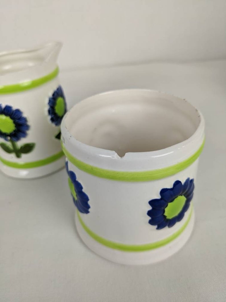 Puede incluir: Un juego de jarra de crema y azucarero de cer&aacute;mica blanca. El juego presenta un dise&ntilde;o floral con flores azules y centros verdes. Bandas verdes rodean la parte superior e inferior de cada pieza. La jarra de crema tiene un pico y un asa.