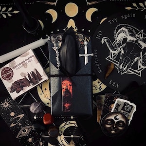 May include: A collection of items including a box of Trumiri Dragon's Blood incense cones, a black wrapped package with a castle bookmark, a silver sword charm, a crystal wand, a white candle, a pendulum, and various celestial-themed cards.